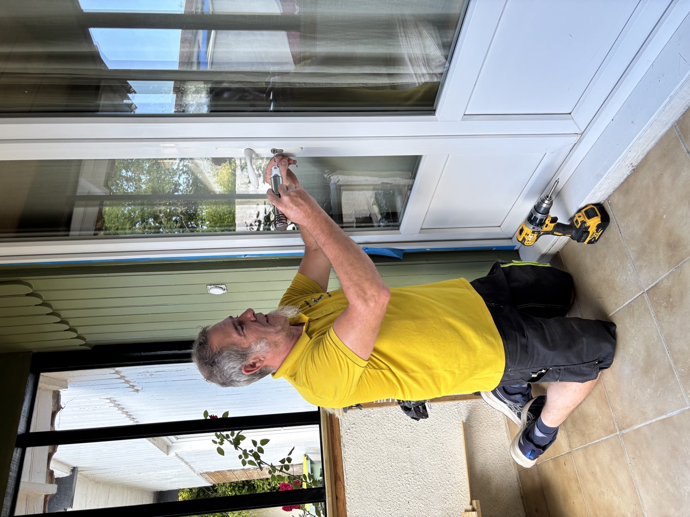 Michel, à genoux, pose une poignée sur une baie vitrée à la perceuse